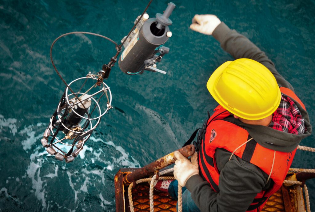 Worker in hard hat and life vest operates machinery on an offshore platform above ocean waters.